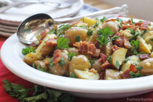 a potato salad dish served on a white ceramic plate. The salad consists of large chunks of cooked potatoes mixed with crispy bacon bits and fresh chopped parsley