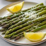 a plate of roasted asparagus topped with grated parmesan cheese and 2 lemon wedges