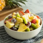 A vibrant fruit salad in a white ceramic bowl placed on a gray and white striped cloth napkin.