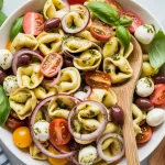 fresh pasta salad in a white ceramic bowl. The salad contains yellow tortellini pasta, halved red and yellow cherry tomatoes, thinly sliced red purple onions, and a chiffonade of fresh green basil leaves