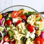 Mediterranean pasta salad photographed from above in a clear glass bowl. The salad contains bow-tie pasta mixed with fresh vegetables including red cherry tomatoes, sliced cucumbers, halved red bell peppers, diced purple onions, and black olives