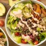 a gourmet salad bowl on a wooden table surface. The bowl contains fresh green lettuce leaves, avocado slices, ground beef, crispy potato wedges, pickle slices, and cherry tomatoes