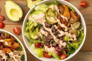 a gourmet salad bowl on a wooden table surface. The bowl contains fresh green lettuce leaves, avocado slices, ground beef, crispy potato wedges, pickle slices, and cherry tomatoes