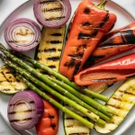 grilled vegetables on a white ceramic plate. The plate contains charred red bell peppers, grilled purple onions in circular slices, bright green asparagus spears